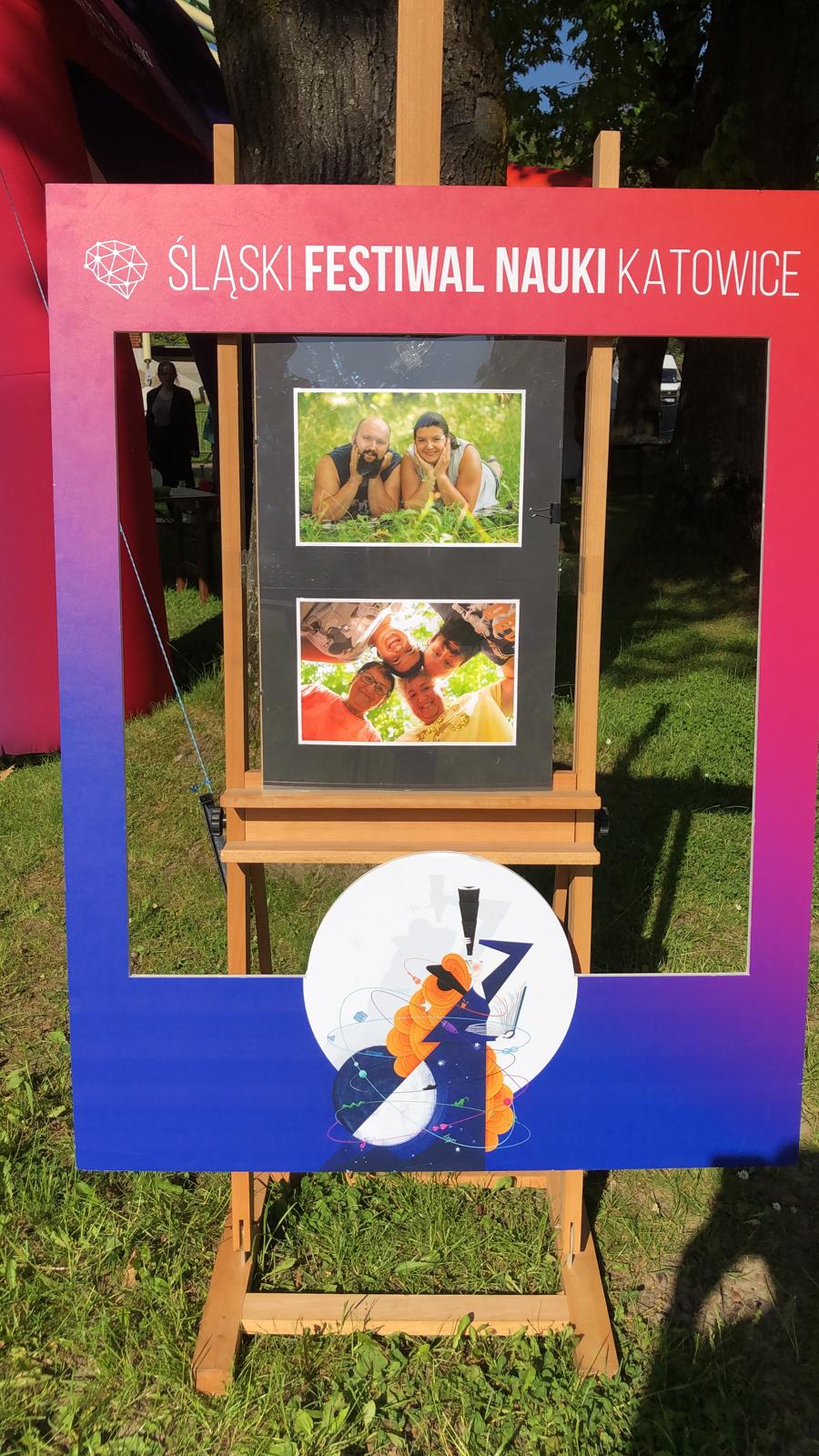 Ramka Sląskiego Festiwalu nauki, w środku sztaluga ze zdjęciami ( 1 zdjecie para leżaca na kocu, drugie zdjęcie rodzina stykajac sie głowami patrzy w dół gdzie jest umieszczony aparat)