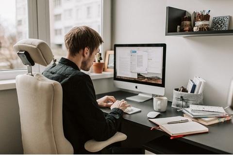 mężczyzna w garniturze siedzi na krześle przy biurku, i pisze na klawiaturze komputera stacjonarnego. Obok niego leżą otwarte notatki.